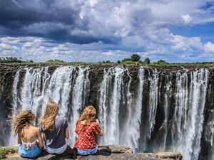Three young people face Victoria Falls while their hair blows in the wind.